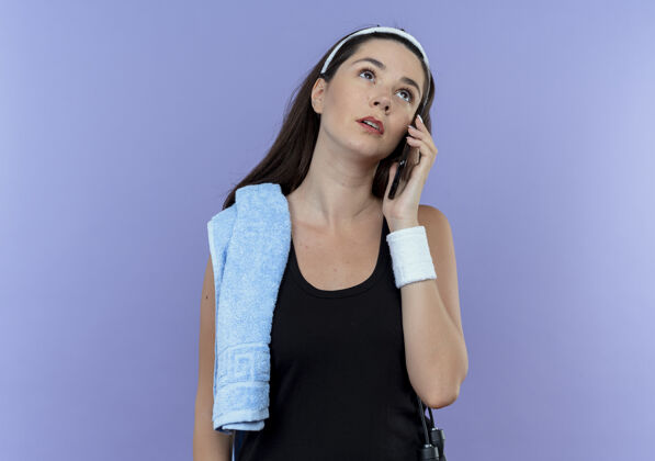 肩膀戴着头巾 肩上扛着毛巾的年轻健身女 站在蓝色的背景上 用沉思的表情看着手机健身支架电话