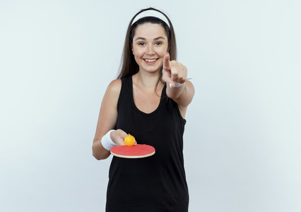桌子戴着头巾 拿着球拍和乒乓球的年轻健身女子 手指对着镜头 站在白色背景上 欢快地微笑着网球年轻人欢呼