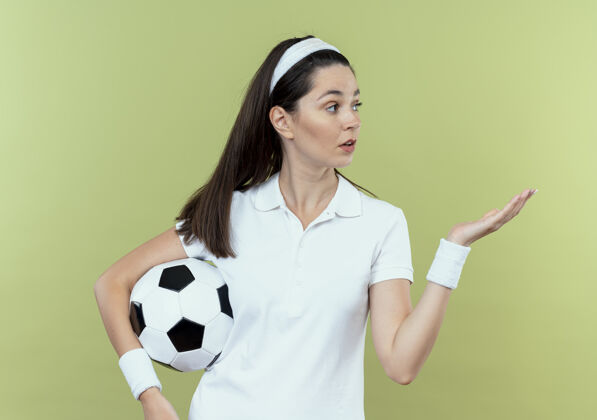 手臂戴着头巾拿着足球的年轻健身女士站在灯光的背景下 手臂看起来很惊讶头带年轻手