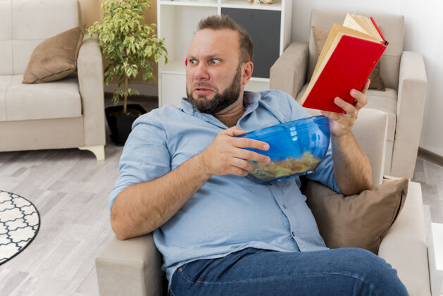 薯片恼怒的成年斯拉夫人坐在扶手椅上 手里拿着一碗薯片和一本书 看着客厅里的一面斯拉夫人碗成人