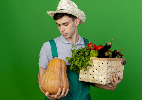 绿色自信的年轻男园丁戴着园艺帽 手里拿着菜篮子 看着隔离在绿色背景上的南瓜年轻复制篮子