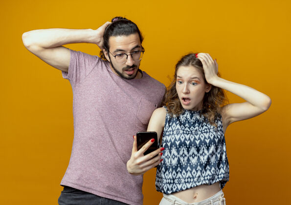 站着站在橙色背景下 一对手持智能手机的年轻夫妇看上去既惊讶又困惑年轻的困惑的情侣