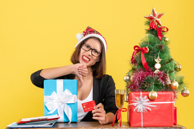 银行迷人的女士穿着西装 戴着圣诞老人的帽子和眼镜 展示着礼物和银行卡 在黄色的办公室里做出消极的手势圣诞老人黄色礼物