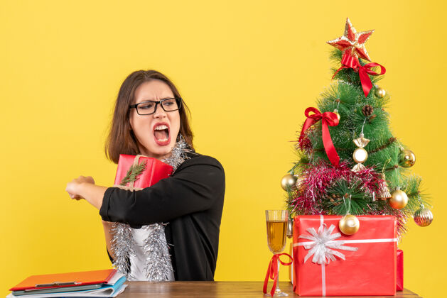 漂亮穿着西装戴着眼镜的商务女士紧张地把礼物藏起来 坐在一张桌子旁 桌子上放着一棵圣诞树坐着礼物成人