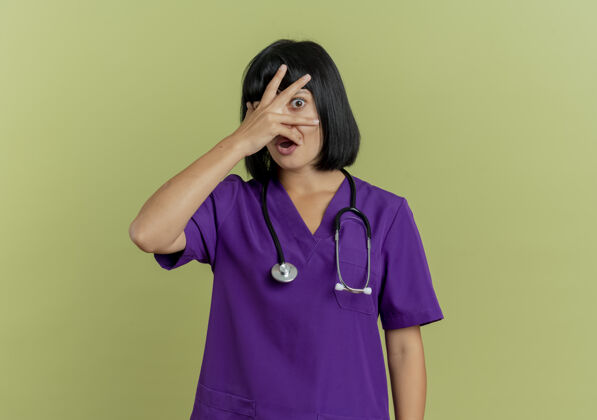 医生害怕的年轻黑发女医生穿着制服 手持听诊器 将手放在脸上 通过橄榄绿背景上的手指观察 并留有复印空间空间复制制服