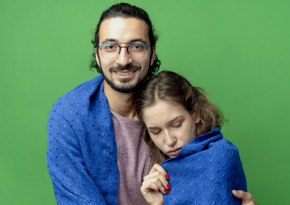 包裹一对年轻的男女 幸福的男人拥抱着他心爱的女朋友 微笑着把她裹在温暖的毯子里 站在绿色的背景上站立毛毯年轻