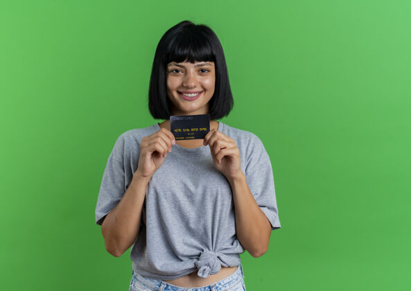 持有微笑着的年轻黑发白人女孩拿着信用卡看着隔离在绿色背景和复印空间的相机空间黑发绿色