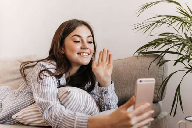 沙发一个穿着条纹衬衫的快乐女人躺在沙发上 通过电话里的视频聊天成人粉色房间