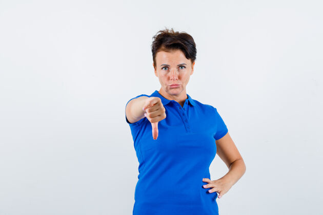 健康成熟女人的肖像 展示拇指在蓝色t恤衫下 看着不高兴的前视图美容柔软提举