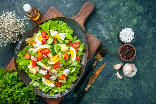 瓶横视美味的沙拉与许多新鲜的原料在木制砧板香料油瓶大蒜素餐具设置在黑绿色混合颜色的背景许多大蒜晚餐