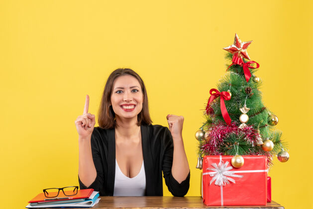 起来一个年轻的女人坐在一张桌子旁 穿着西装骄傲地指着黄色的办公室装饰的圣诞树漂亮骄傲成人