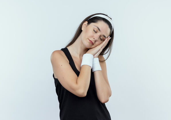 手掌戴着头巾的年轻健身女士做着睡觉的姿势 手掌靠在手掌上 闭着眼睛站在白色的背景上年轻眼睛睡眠