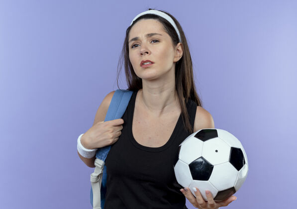 球戴着头巾 背着背包 拿着足球的年轻健身女士站在蓝色背景下 看上去很困惑背包困惑健身