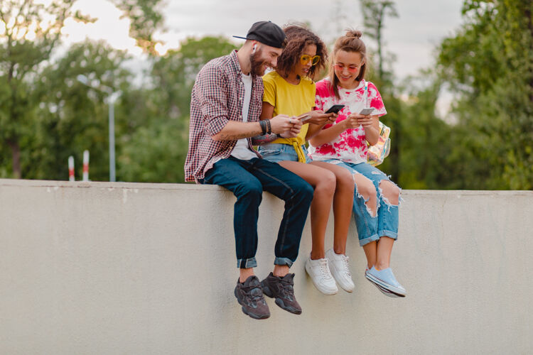 音乐快乐的年轻朋友坐在公园里用智能手机微笑公司享受兴奋手机