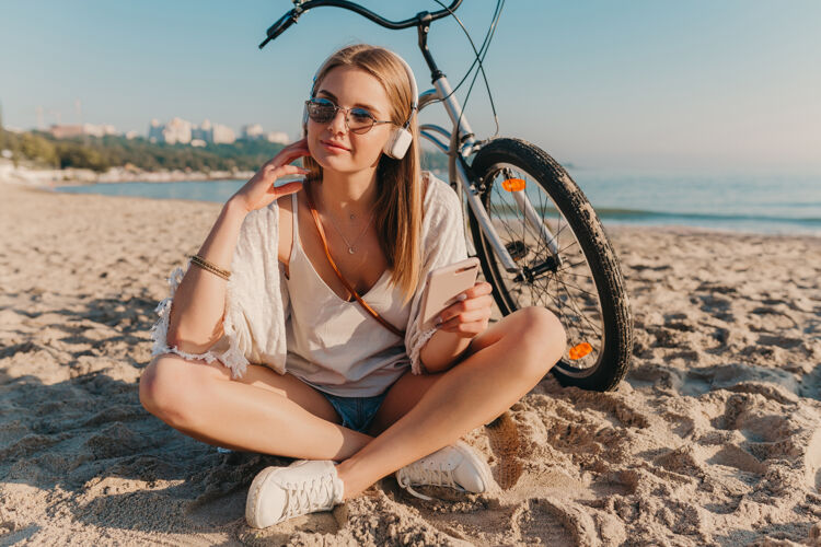 休闲时尚年轻迷人的金发微笑的女人坐在沙滩上骑着自行车戴着耳机听音乐海滩海岸积极