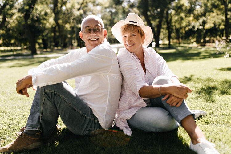 成人积极的女士戴着凉爽的帽子 穿着条纹时髦的衬衫和牛仔裤 微笑着坐在草地上 男士戴着眼镜 在户外穿着轻便的衬衫情侣退休拥抱