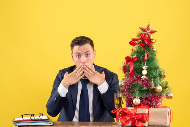 管乐器前视图惊奇的男人把手放在嘴边 坐在圣诞树旁的桌子上 呈现黄色背景男礼物惊奇的人