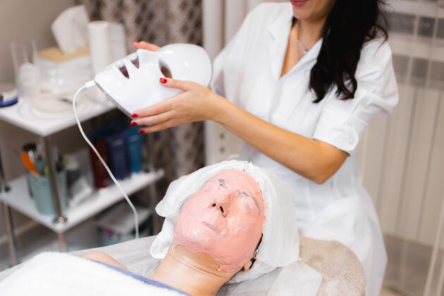 美容师客户躺在美容桌上的沙龙里 脸上戴着白色的面具面部皮肤科女性