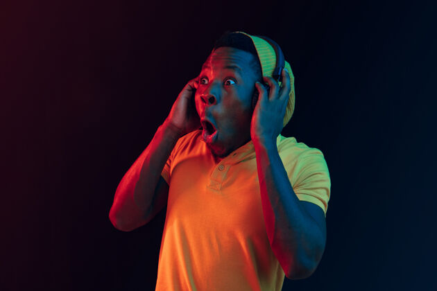 牙买加年轻帅气快乐的时髦男人在霓虹灯下用耳机在黑工作室听音乐迪斯科舞厅 夜总会 嘻哈风格 积极的情绪 面部表情 舞蹈概念成人脸帽子