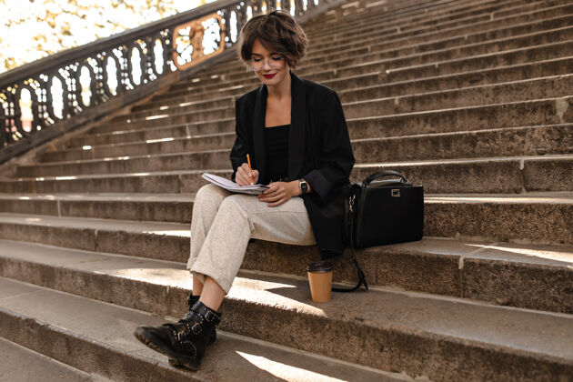 户外穿着轻便裤子 黑色夹克和靴子的时髦女人坐在外面的楼梯上戴眼镜的短发女人在户外写字包城镇走道