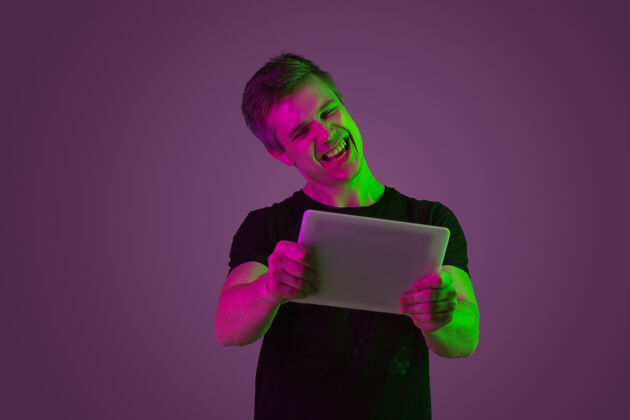 情绪玩平板电脑 欣赏霓虹灯下紫色工作室背景上的白种人肖像穿着黑色衬衫的漂亮男模人类情感的概念 面部表情 销售 广告面部帅气员工