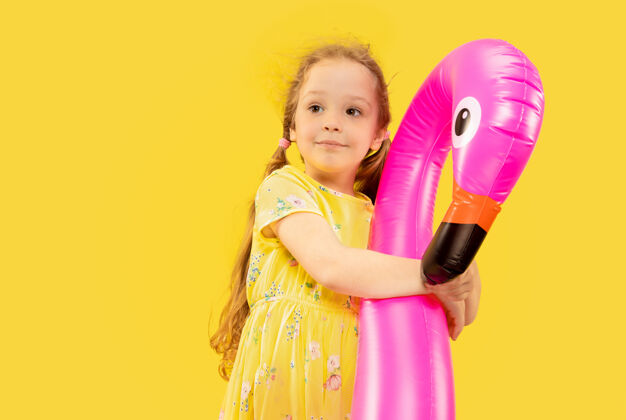 火烈鸟美丽动情的小女孩被隔离在黄色背景上半幅快乐的孩子的肖像 穿着裙子 手里拿着橡胶粉色的火烈鸟夏天的概念 人类的情感 童年手势孩子橡胶