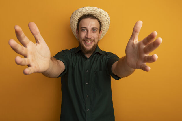 手微笑的金发帅哥 戴着沙滩帽 伸出双手孤立在橙色的墙上伸展微笑人