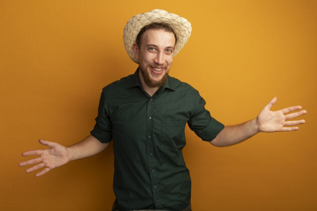 站快乐的金发帅哥戴着沙滩帽 在橙色的墙上张开双手开帅哥人