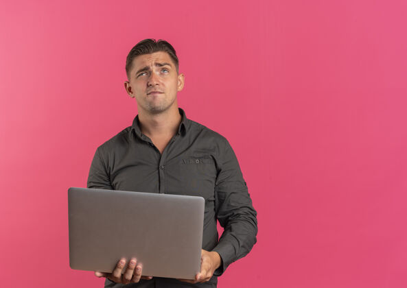 笔记本电脑年轻体贴的金发帅哥拿着笔记本电脑 在粉色背景上孤立地抬头看 还有复印空间英俊金发年轻