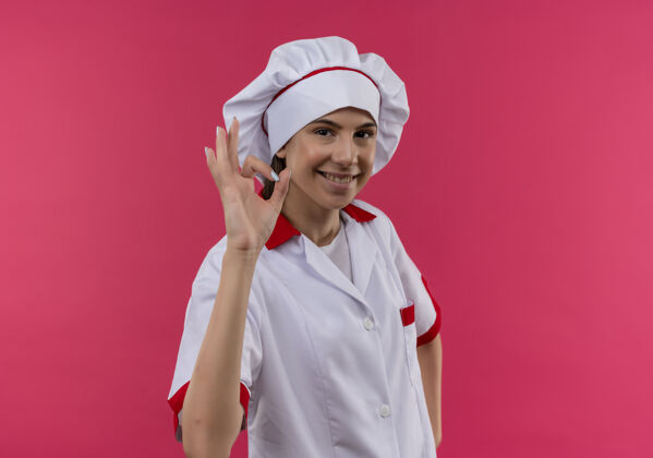 好年轻微笑的白人厨师女孩在厨师制服手势ok手势孤立的粉红色背景与复制空间手势年轻微笑