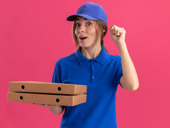 制服穿着制服的年轻漂亮的送货员兴奋地举起拳头 把比萨饼盒孤立地放在粉红色的墙上表情姿势盒子