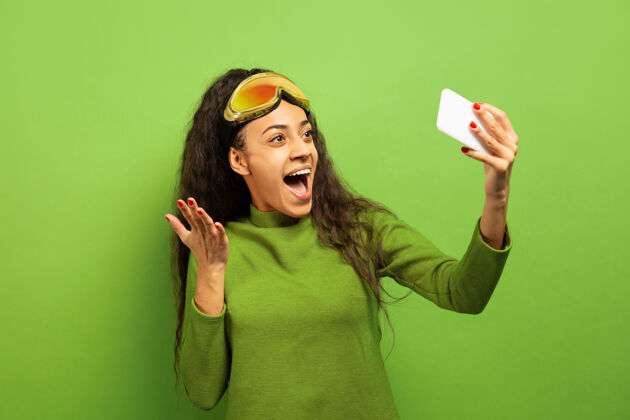 肖像以绿色工作室为背景 黑色皮肤的非洲裔美国年轻女子滑雪面具肖像关于人类情感 面部表情 销售 广告 冬季运动和假期的概念自拍或虚拟博客成人女孩美国