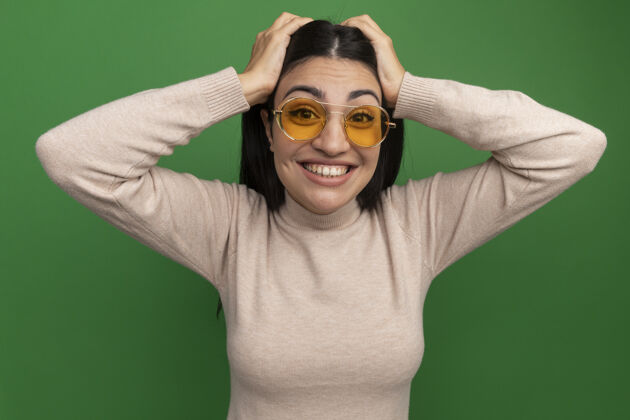 眼镜兴奋的黑发美女戴着太阳镜把手放在头上隔离在绿色的墙上兴奋市民脸
