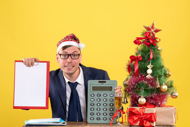 桌子正面图：一个戴着圣诞帽的男人坐在圣诞树旁的桌子旁 黄色背景的礼物圣诞节圣诞老人职业