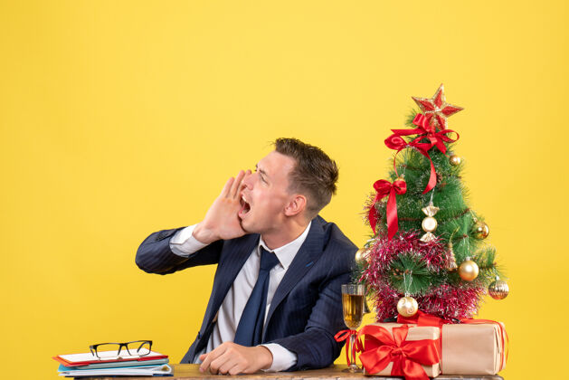 男前视图愤怒的男人叫某人坐在圣诞树旁的桌子上 黄色背景上的礼物前面行政新郎