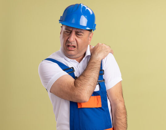 脸疼痛的成年建筑工人穿着制服 把手放在肩膀上 隔离在橄榄绿的墙上人手人