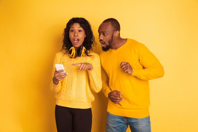 微笑年轻感性的非裔美国人 穿着黄色背景的明亮休闲服美丽的情侣人类情感的概念 面部表情 关系在智能手机上震惊地指着男性非洲化妆品