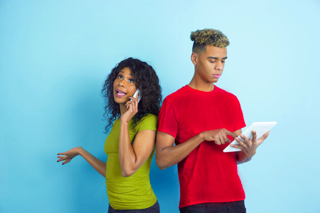 关系在平板电脑上工作 在电话上交谈年轻情绪化的非裔美国人男女在蓝色背景上穿着五颜六色的衣服美丽的情侣人类情感的概念 面部表情 关系 广告化妆品情侣非洲