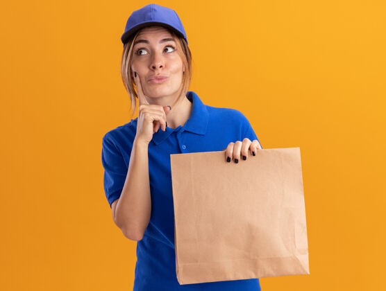 包装穿着制服的年轻漂亮的女送货员把手指放在脸上 拿着纸包 看着橘色墙上孤立的一面年轻姿势立场
