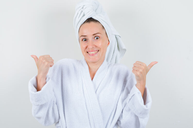 健康身穿白色浴袍的年轻女子 毛巾上竖起大拇指 看上去很开心 正前方的景色肖像毛巾乐趣
