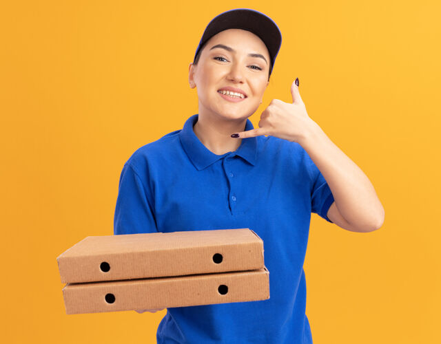 站着身穿蓝色制服 头戴鸭舌帽的年轻女送货员拿着比萨饼盒 面带微笑地站在橙色的墙上 摆出“叫我”的手势手势表演欢呼