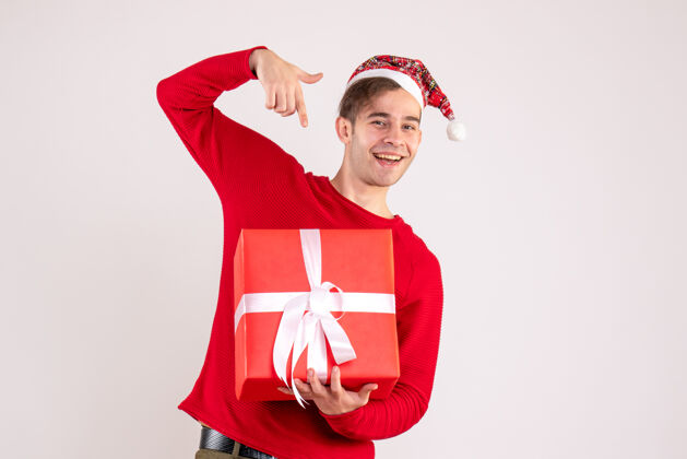 微笑前视图：戴圣诞帽的年轻人指着白色背景上的礼物可爱帽子快乐