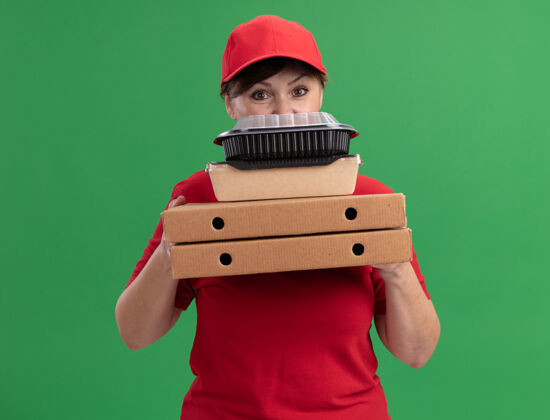 女人快乐的中年女送货员穿着红色制服 戴着帽子 手里拿着披萨盒和食品包 站在绿色的墙上看着前面食物披萨盒子