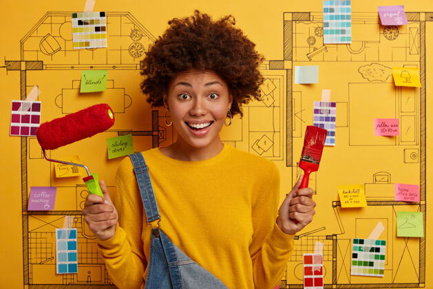 样品正忙的女油漆工拿着油漆辊和刷子 做房屋维修 穿着黄色的套头衫和工作服素描刷子非洲