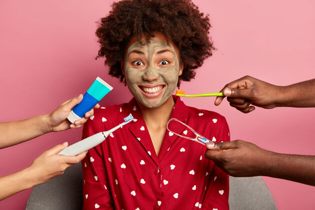 坐着积极黑皮肤的年轻女子涂上粘土面膜 有着牙齿般的微笑 关心牙齿 双手围着牙刷 牙膏化妆洗脸皮肤护理