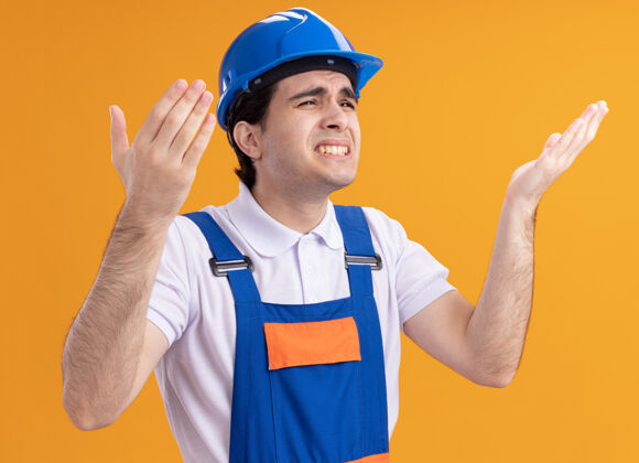 站着身穿施工制服 头戴安全帽的年轻建筑工人站在橙色的墙上 抬头望去 感到困惑和不快不高兴男人建筑