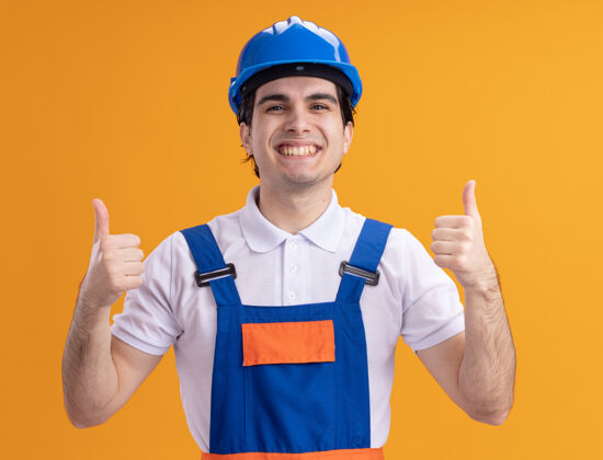 欢呼年轻的建筑工人身穿建筑制服 戴着安全帽 站在橙色的墙上 面带微笑 高高兴兴地竖起大拇指表演拇指安全