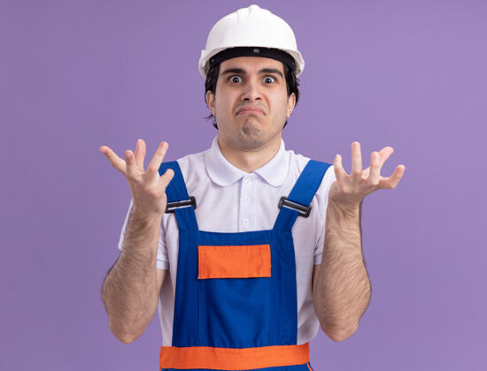 困惑年轻的建筑工人穿着建筑制服 戴着安全帽 困惑和惊讶地举起双臂看着前面站在紫色的墙上安全建筑男人