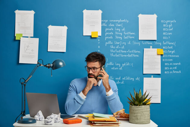 台灯穿着蓝色套装的忙碌的男性自由职业者 检查笔记本电脑上的信息 使用现代手机小玩意打电话 坐在白纸桌旁纸张自由职业者教育