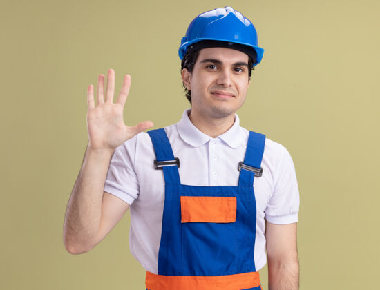 脸年轻的建筑工人穿着建筑制服 戴着安全帽 面带微笑 站在绿色的墙上挥手致意安全微笑男人